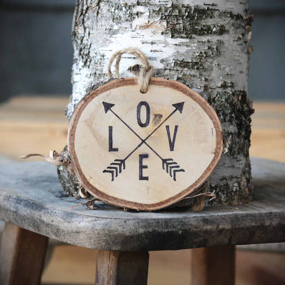 A rustic round birch wood ornament leaning on a tree trunk, featuring the word LOVE spelled out with crossed arrows and arrow details in black on a light natural wood background.