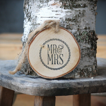 A round natural birch wood slice ornament with a rustic hanging twine and a black stamped 'MR & MRS' monogram, displayed against a birch tree trunk on a wooden stool.