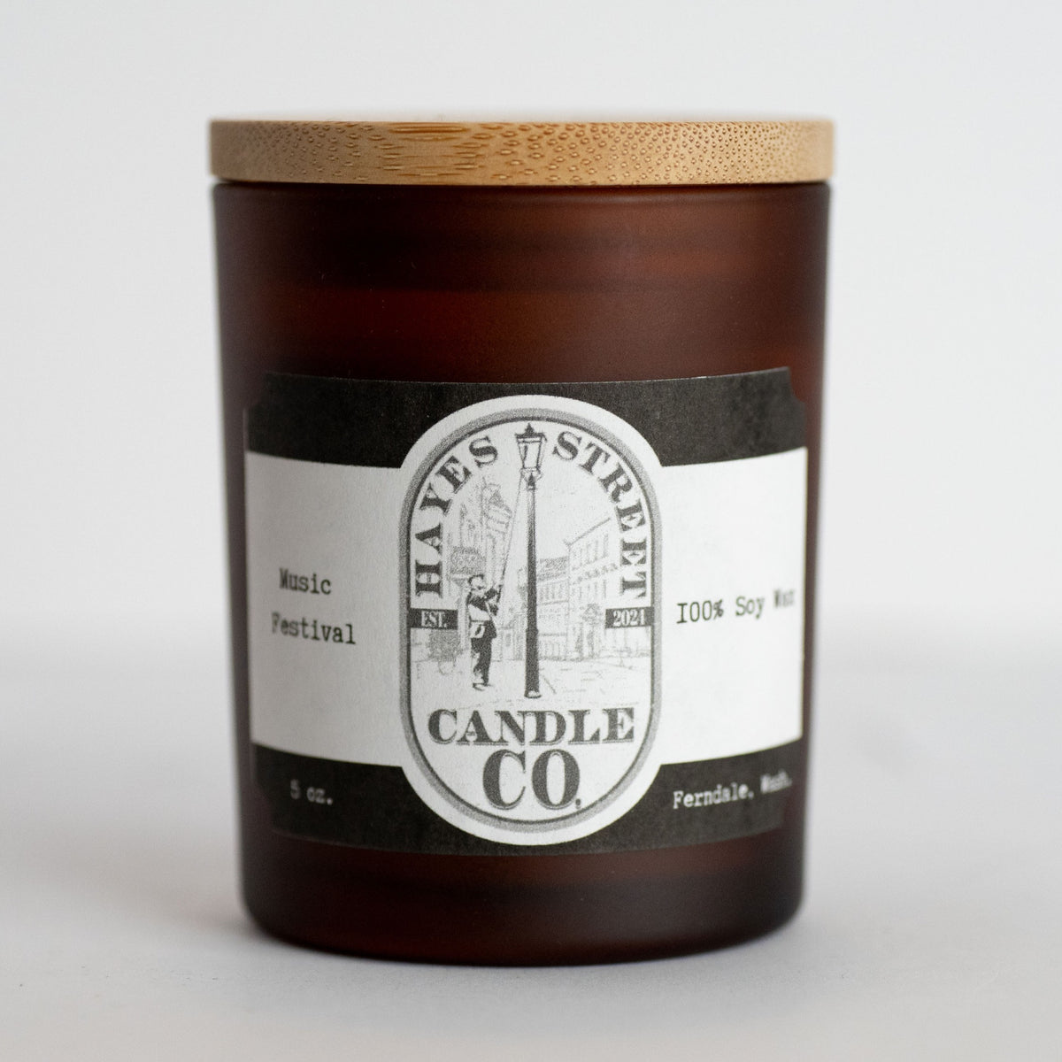 An amber glass jar candle with a light wood bamboo lid and a black and white paper label reading 'Hayes Street Candle Co.' on a clean white background. The scent is Music Festival and it is made in Ferndale, Wash.