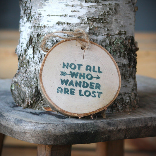 A round natural wood slice ornament with the phrase 'NOT ALL WHO WANDER ARE LOST' in dark grey lettering, hanging from a twine loop in front of a birch tree trunk on a rustic wooden stool. It is offered by Hayes Street Candle Company in Ferndale, Washington.