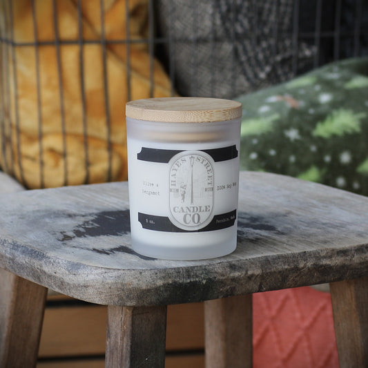 A frosted glass candle jar with a light wood lid sits on a rustic wooden stool. The label reads Hayes Street Candle Company and Olive and Bergamot. Ferndale, WA