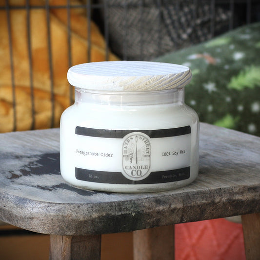 A jar candle labeled 'Pomegranate Cider' by Hayes Street Candle Co., sitting on a rustic wooden stool with a blurred indoor background.