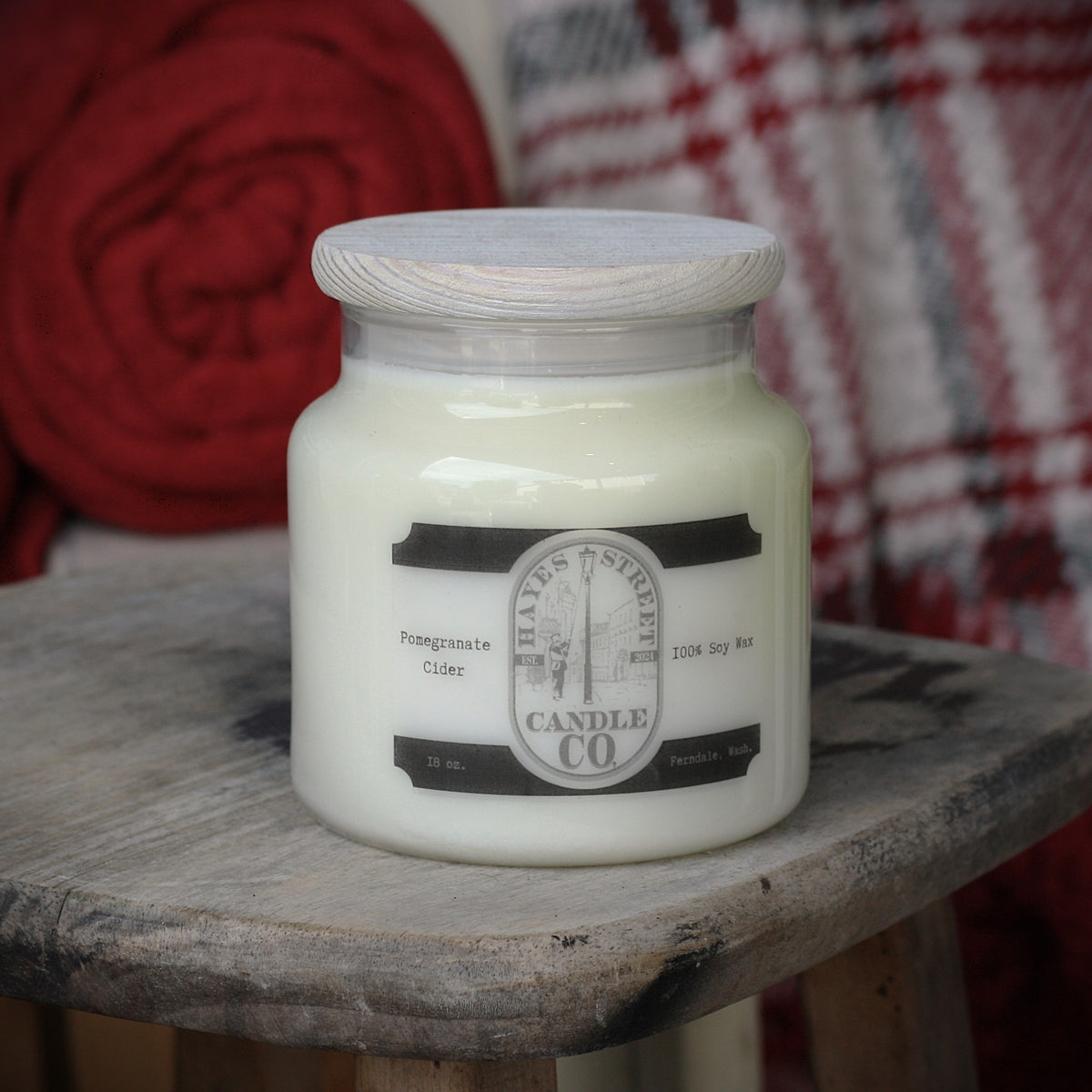 A jar candle with white wax sits on a rustic wooden stool. The candle has a light wood lid and a label that reads Hayes Street Candle Company with a little man lighting a street lamp. The scent is Pomegranate Cider and is made in Ferndale, Washington.