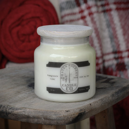 A jar candle with white wax sits on a rustic wooden stool. The candle has a light wood lid and a label that reads Hayes Street Candle Company with a little man lighting a street lamp. The scent is Pomegranate Cider and is made in Ferndale, Washington.