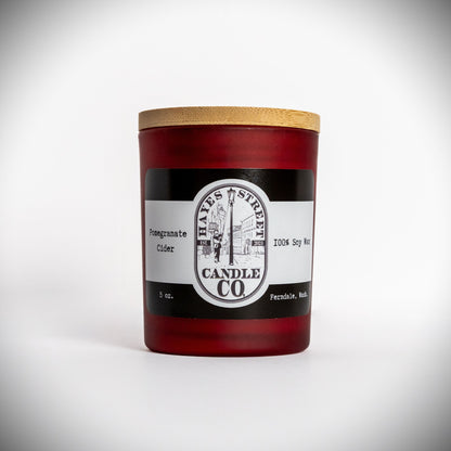A 5 oz soy wax candle in a dark red glass jar with a light wood lid, featuring a black and white label from Hayes Street Candle Co. on a white background. The scent is Pomegranate Cider