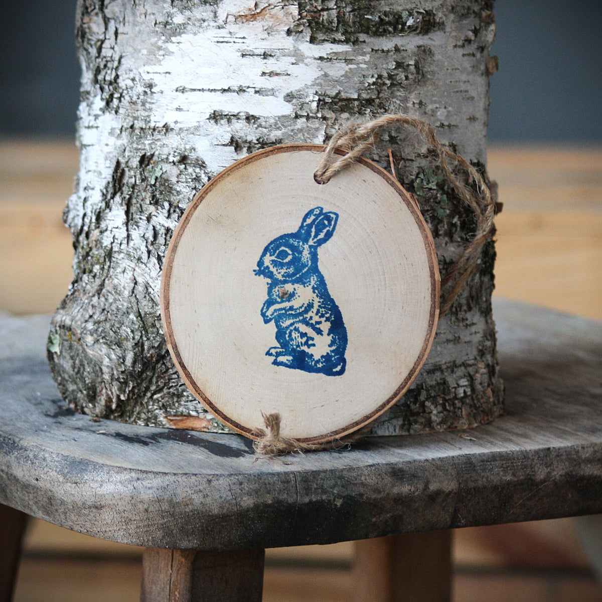 A round wooden slice ornament with a blue rabbit print and twine hanger, leaning against a birch tree on a rustic wooden stool.