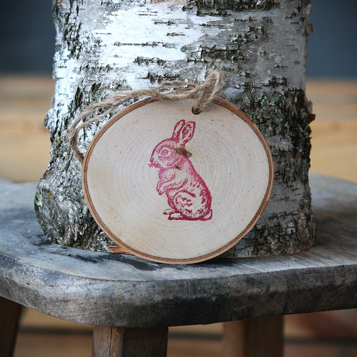 A round birch wood slice ornament with a red stamped rabbit design, hanging from a twine loop in front of a birch tree trunk on a rustic wooden stool. Made from Log Slices by Margie and Hayes Street Candle Company of Ferndale, Washington.