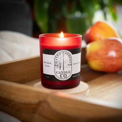 A lit red glass candle jar labeled 'Hayes Street Candle Co.' sits on a wooden tray, with apples blurred in the background. The scent is Red Apple Bliss.