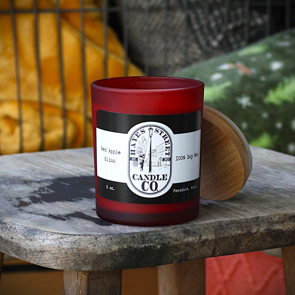 A red frosted glass candle jar with a black and white label reading 'Hayes Street Candle Co' and 'Red Apple Bliss', sitting on a rustic wooden table with its wooden lid leaning against it. The background shows a cozy indoor setting with a yellow blanket and patterned cushions.