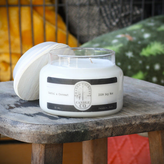 A white soy wax candle in a clear glass jar with a white lid resting beside it on a rustic wooden stool. The label reads 'Santal & Coconut' and '100% Soy Wax' with the brand Hayes Street Candle Co.