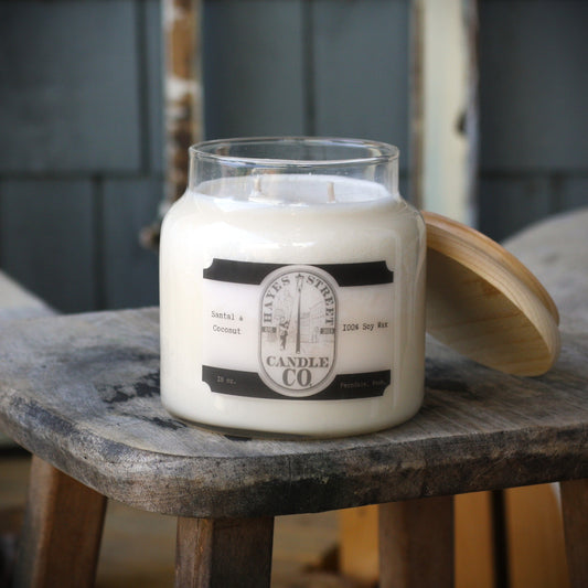 A jar candle with a clear glass container and white wax sits on a rustic wooden stool, with its wooden lid leaning against the right side. The front label reads 'HAYES STREET CANDLE CO' and 'Santal & Coconut'.
