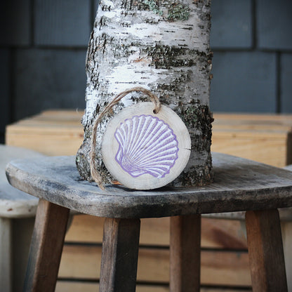A round wooden hanging ornament with a purple seashell design, suspended by a twine loop from a birch tree trunk on a rustic wooden stool.
