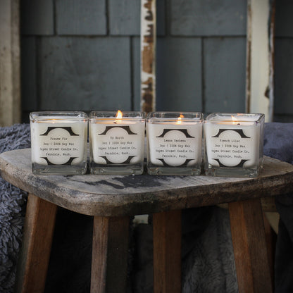 Four lit soy candles in clear square glass jars arranged on a rustic wooden stool, with a dark gray shingled wall in the background.