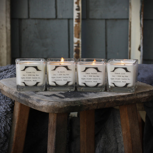 Four lit soy candles in clear square glass jars arranged on a rustic wooden stool, with a dark gray shingled wall in the background.
