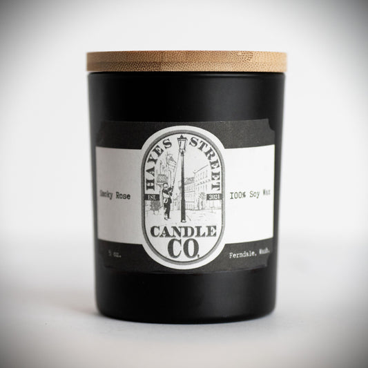 A black glass jar candle with a light wood bamboo lid and a white and black label reading Hayes Street Candle Co. and 'Smoky Rose 100% Soy Wax'.
