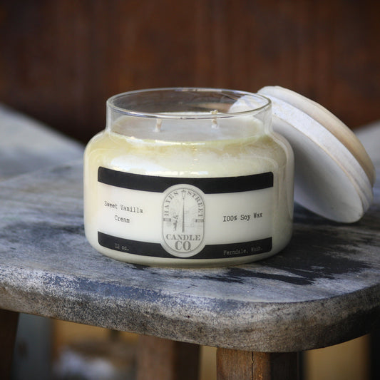Sweet Vanilla Cream candle from Hayes Street Candle Company. It's 100% soy wax (which is what it reads on the label) and is made in Ferndale, Washington. The candle pictured is a double wick in an apothecary style jar, with a white lid leaning against it. The candle rests on a rustic wooden stool, with a dark rusted brown behind it.