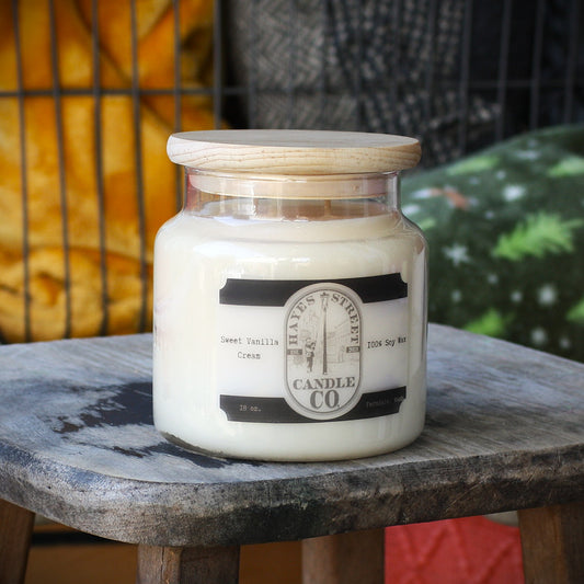 A jar candle labeled 'Sweet Vanilla Cream' by 'Hayes Street Candle Co.' sits on a rustic wooden stool.