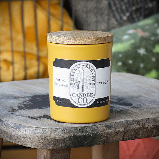 A yellow candle jar with a bamboo wood lid sits on a weathered wooden table. The label reads 'Hayes Street Candle Co' and 'Tropical Fruit Punch'.