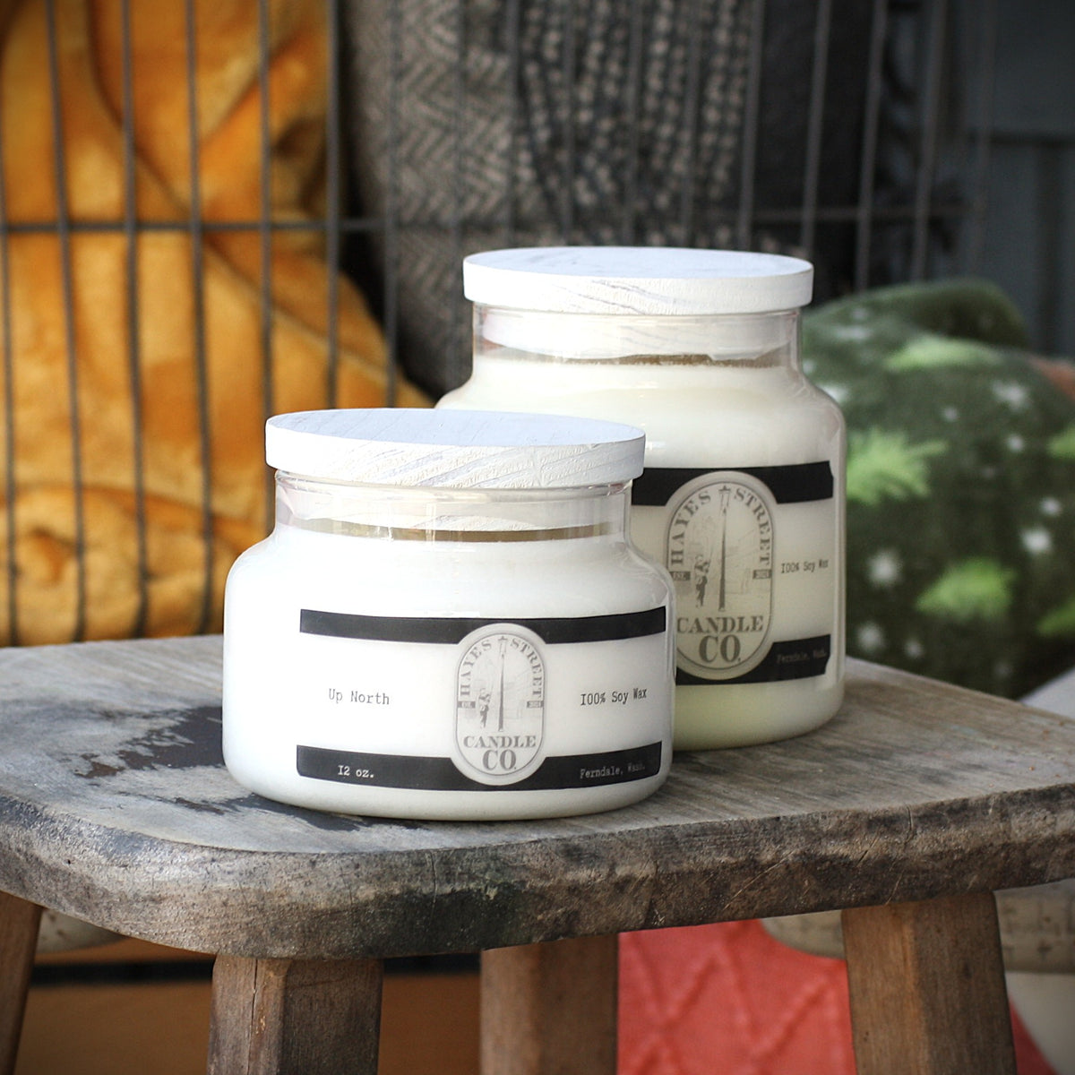 Two glass soy wax candles with white lids and black-and-white labels from Hayes Street Candle Co., displayed on a rustic wooden stool with a cozy indoor background.