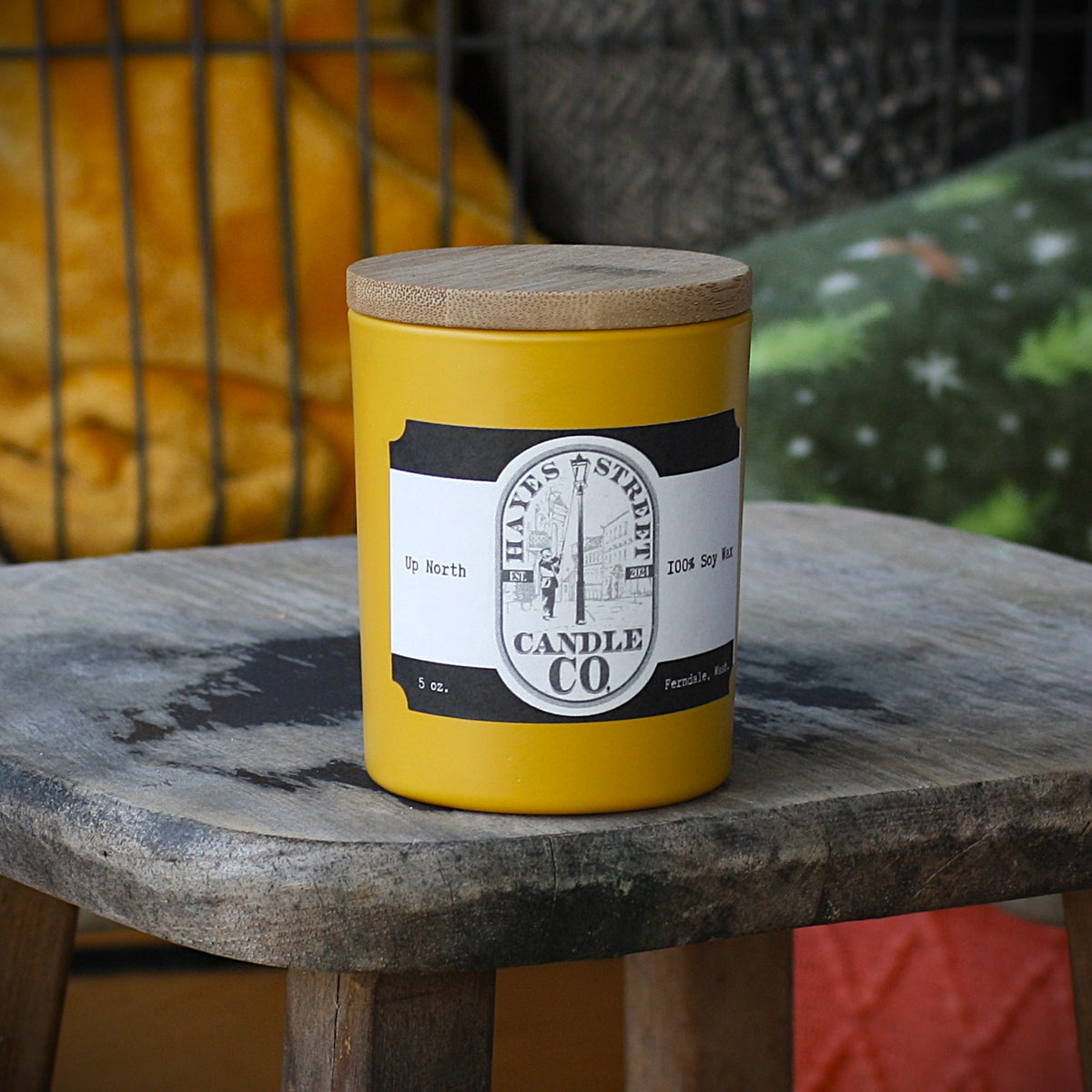 A yellow jar candle with a wooden lid sits on a rustic wooden stool. The candle features a white and black label with the brand name 'HAYES STREET CANDLE CO.