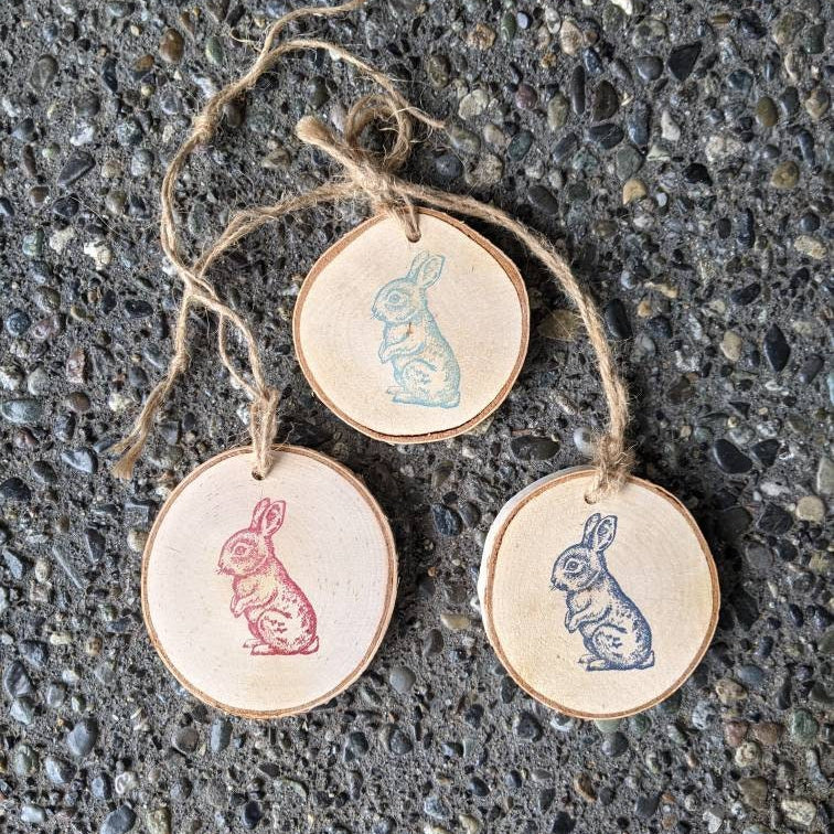 Three round wooden ornaments with rabbit illustrations hanging on a string against a textured surface.